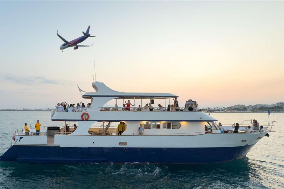 A white and blue two-deck tour boat carries passengers off the Larnaca coast at dusk while a passenger jet flies overhead