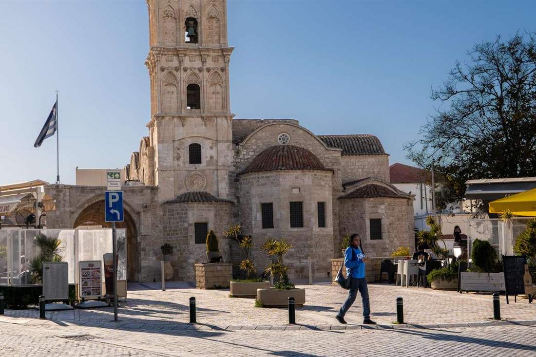 Stone bell tower and domed apses of the Church of Saint Lazarus in Larnaca under a blue sky, with a pedestrian crossing the cobbled square