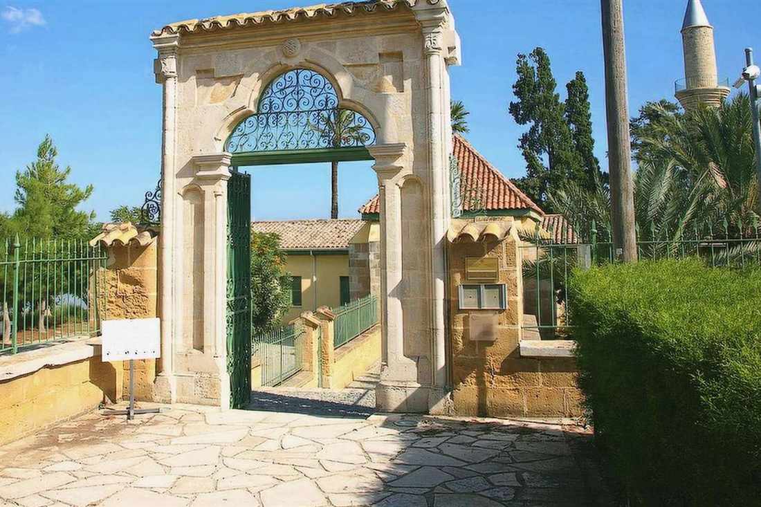 Carved stone gateway with a wrought-iron arch and open green gate, with a minaret and palm trees behind it