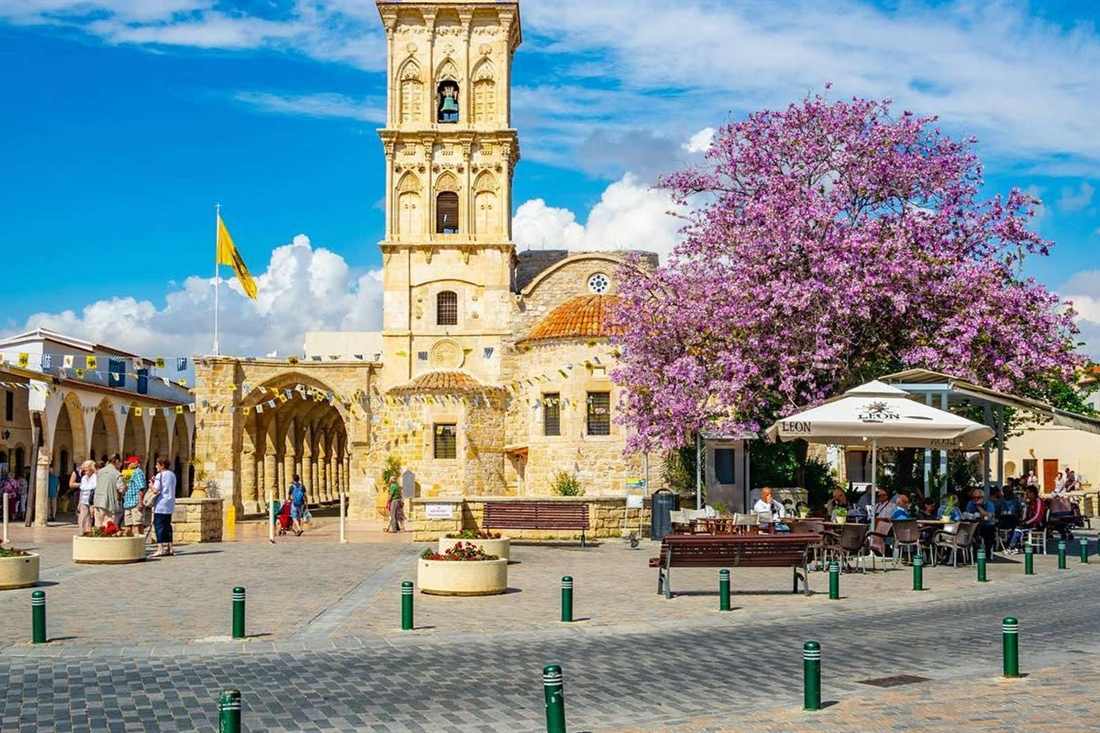 Bell tower and stone arcade of the Church of Saint Lazarus in Larnaca beside a flowering purple tree under a blue sky with white clouds