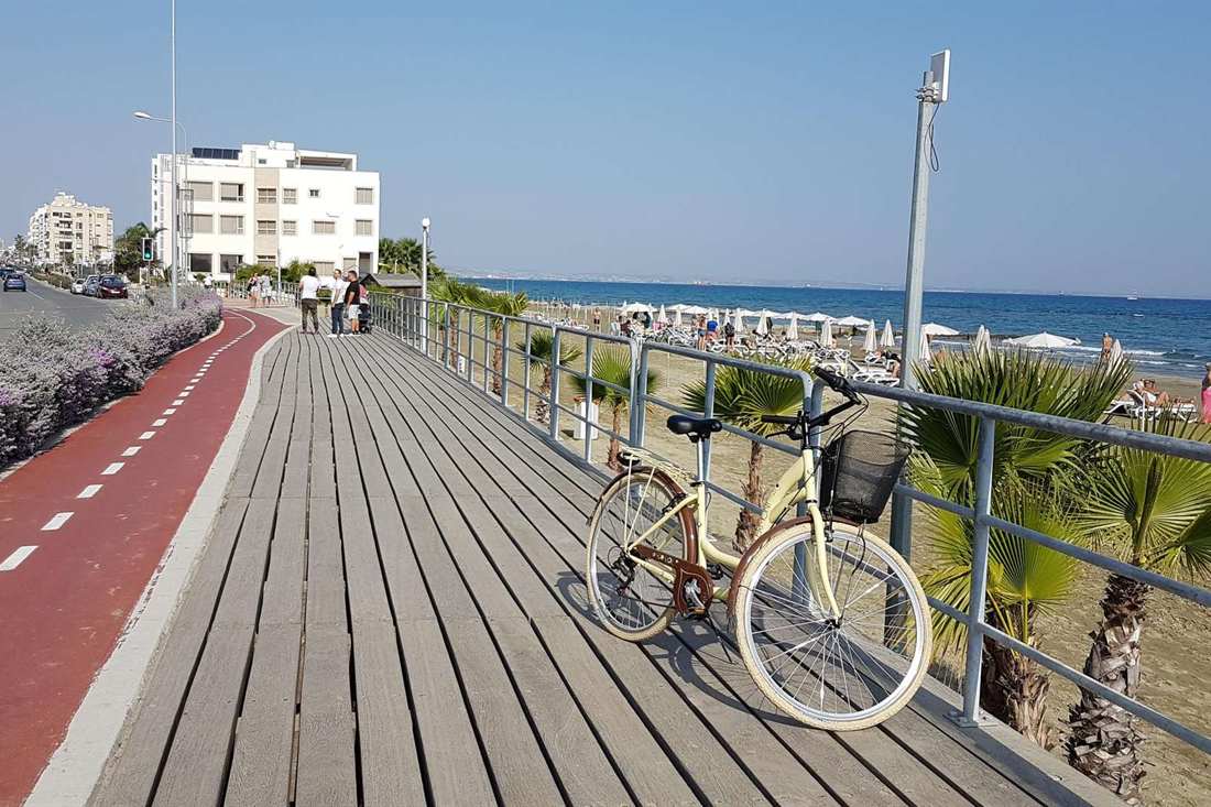 A cream city bicycle with a front basket parked against the railing of the Larnaca seafront boardwalk beside a red cycle lane, with the sea and beach umbrellas behind