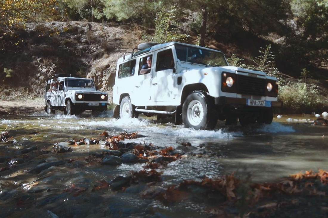 Two white Land Rover Defender jeeps driving through a shallow stream between rocks and autumn leaves in a forest