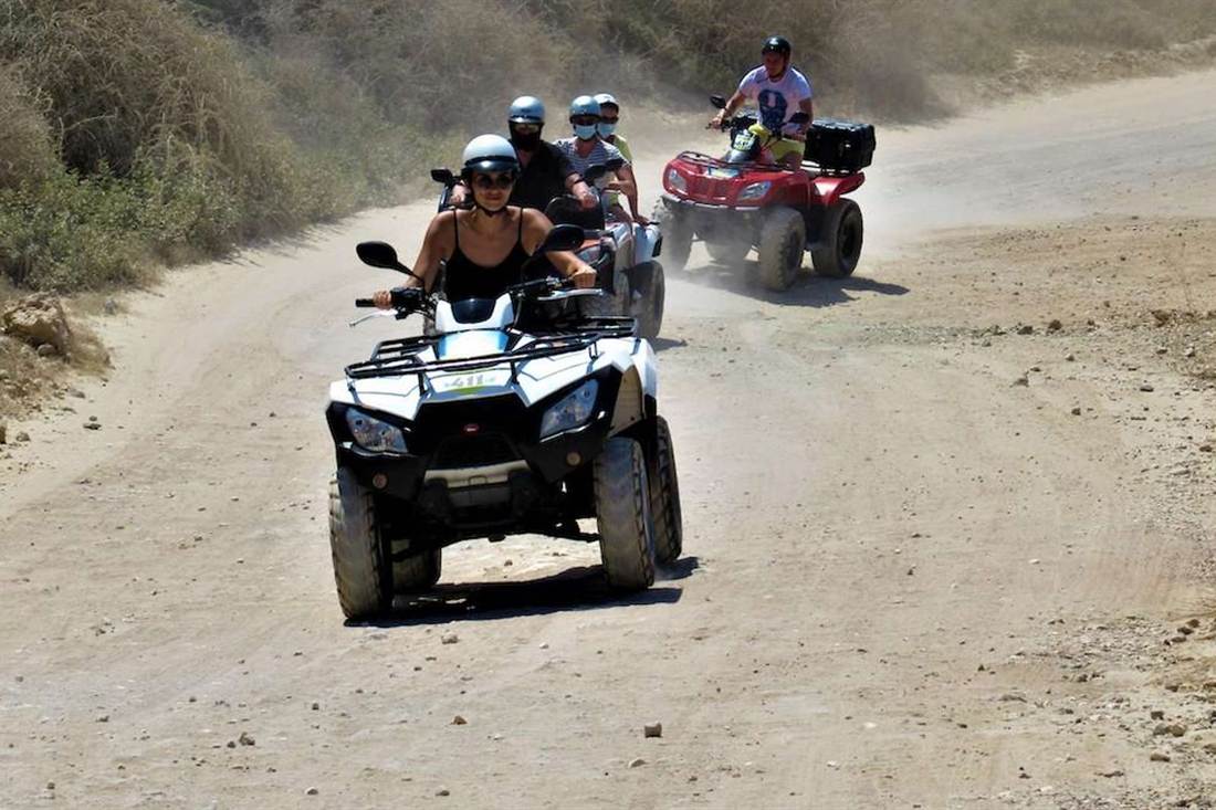 A rider in a white helmet drives a white quad bike along a dusty dirt track, with more riders on quads behind her