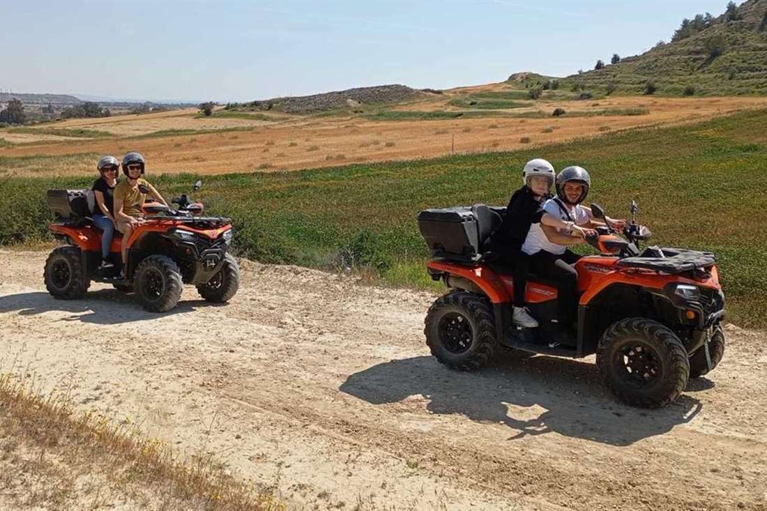 Two orange quad bikes with riders and passengers on a dirt track beside green and golden fields and low hills