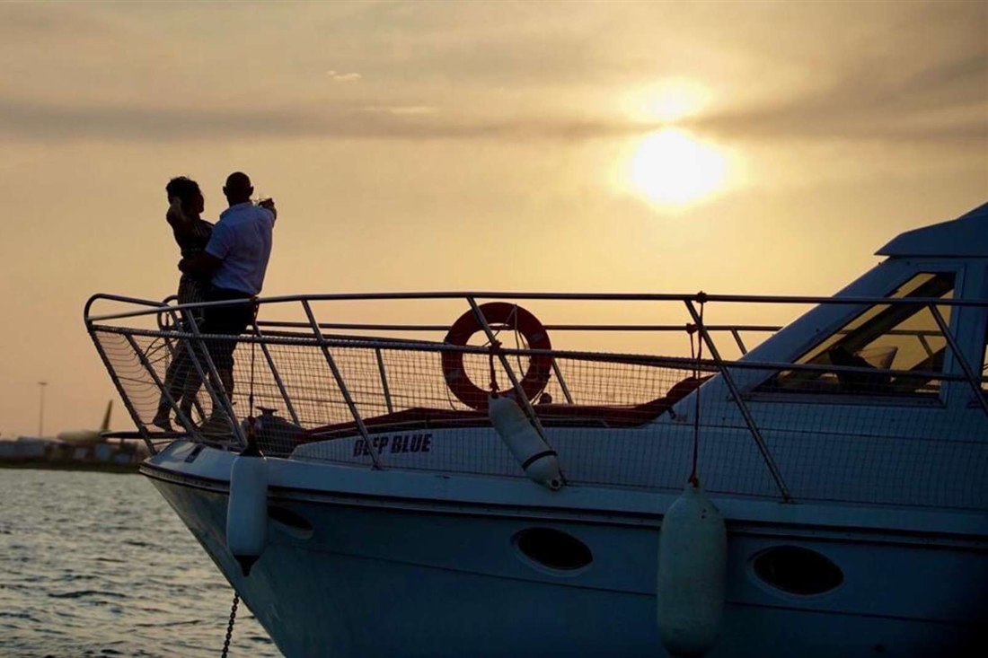 A white yacht at golden hour with two people silhouetted at the bow rail and the low sun behind soft cloud over the sea