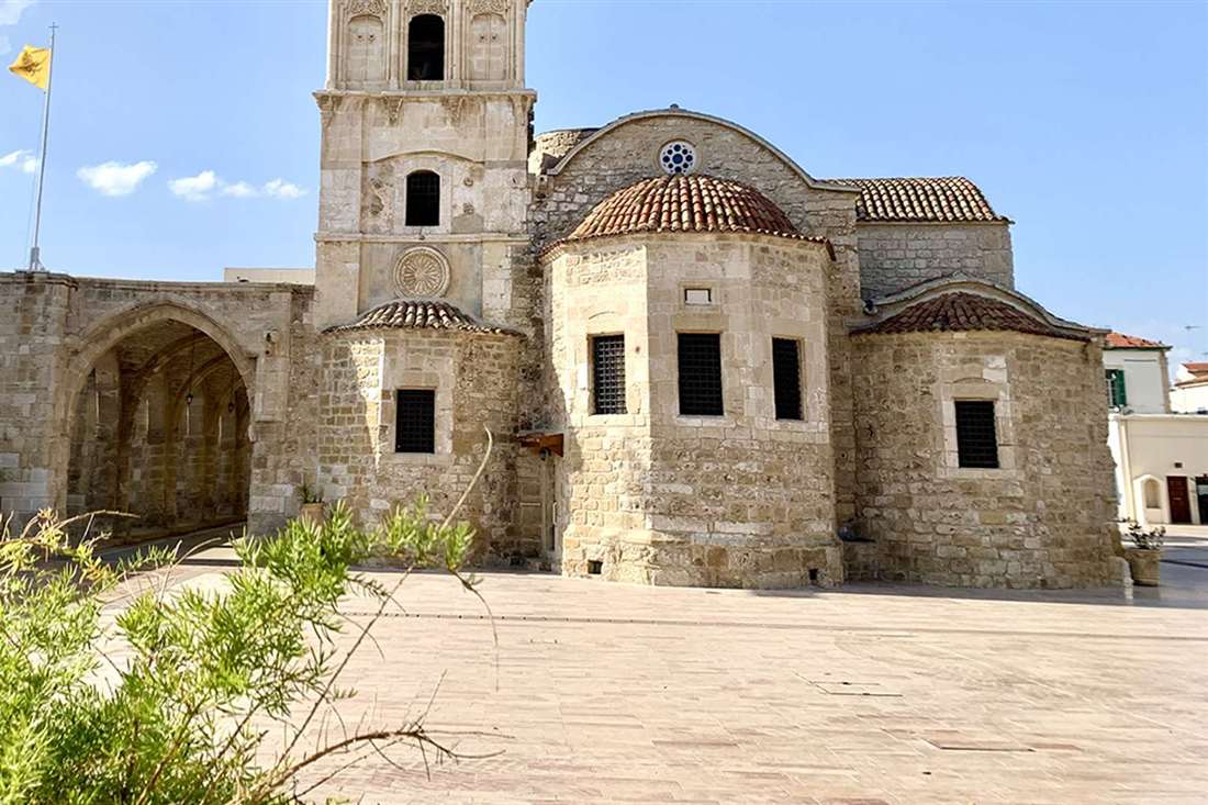 Stone Church of Saint Lazarus in Larnaca with its bell tower, red-tiled domes and a paved square under a blue sky
