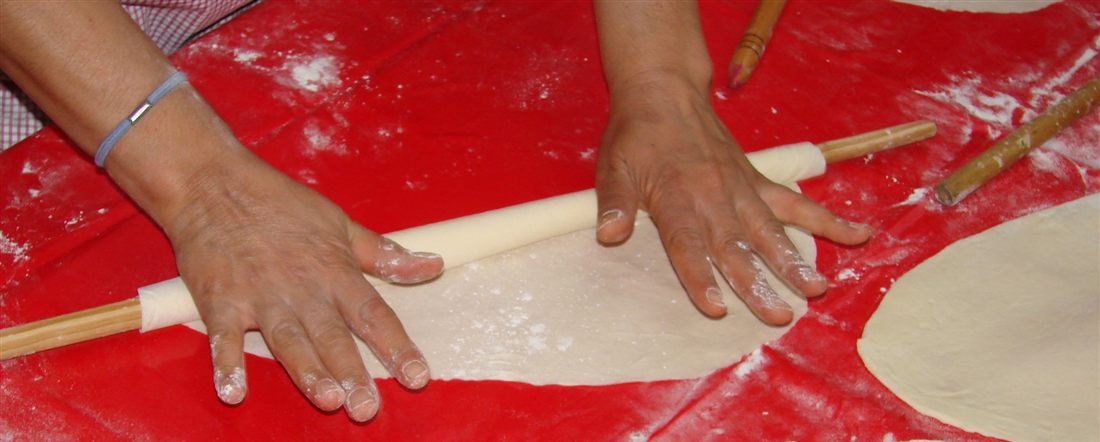 Hands rolling thin pastry sheets on a floured table with a long wooden rolling pin, the core skill of a phyllo pie Lefkada cooking class.