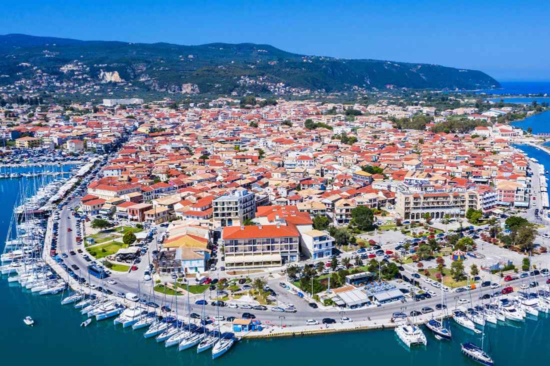 Aerial view of Lefkada Town with red-roofed houses, a marina full of sailboats along the waterfront and green hills behind under a clear blue sky
