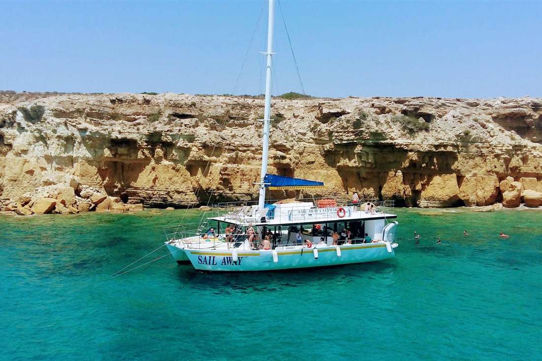 White catamaran Sail Away at anchor in turquoise water below sandstone cliffs, with swimmers beside the boat