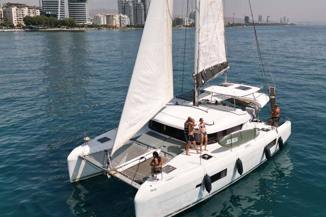 White sailing catamaran with both sails up and people on deck, cruising on blue sea off the Limassol seafront with apartment towers on the shore