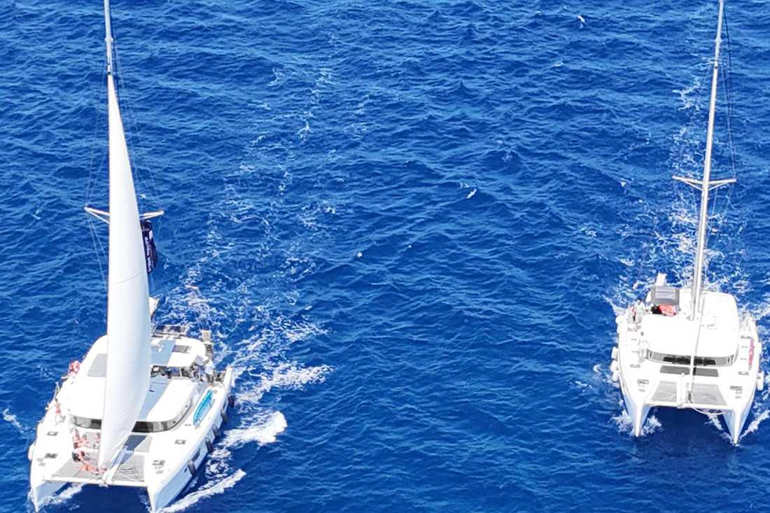 Two white sailing catamarans cruising side by side on deep blue open sea, seen from above