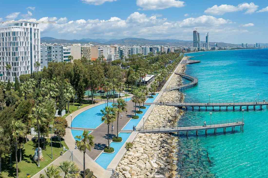 Aerial view of the Limassol seafront promenade with palm trees, blue pools, wooden piers over turquoise sea and high-rise buildings along the coast under a blue sky