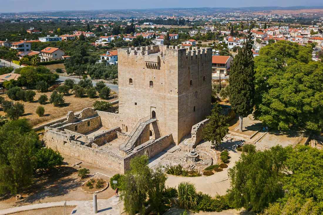 Aerial view of the square stone keep of Kolossi Castle with battlements and an outer staircase, surrounded by green trees and village houses on a sunny day