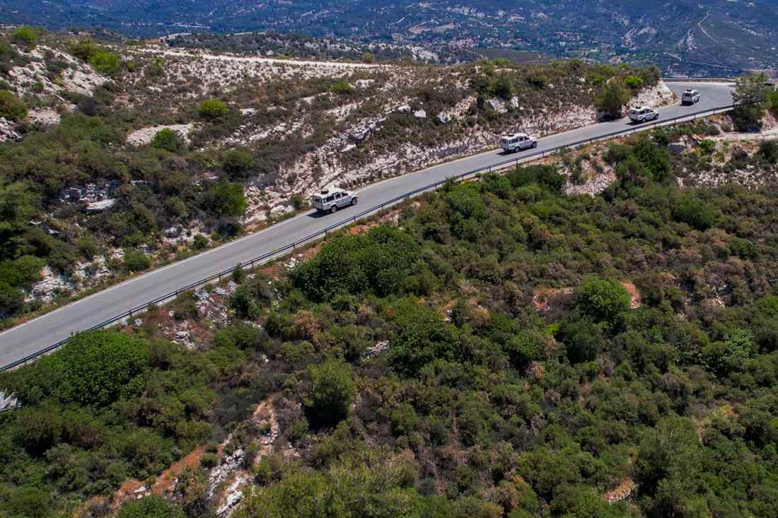 A line of white jeeps on a winding mountain road through green scrub and white rock, seen from above