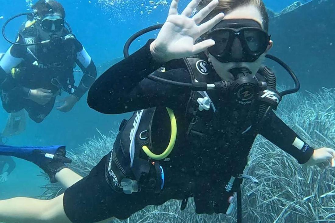 Scuba diver in a black wetsuit waving at the camera underwater, with another diver behind, over a seagrass seabed in clear blue water