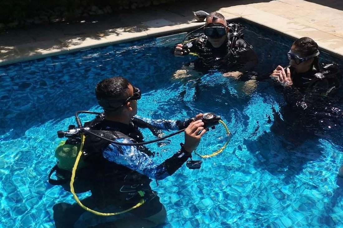 Scuba instructor holding a regulator and gauge in a blue tiled swimming pool while two divers in masks and wetsuits watch