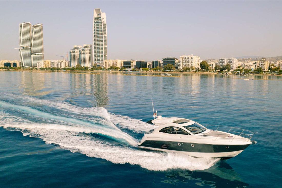 White motor yacht speeding across calm blue water off the Limassol seafront, with high-rise towers and the coast behind it