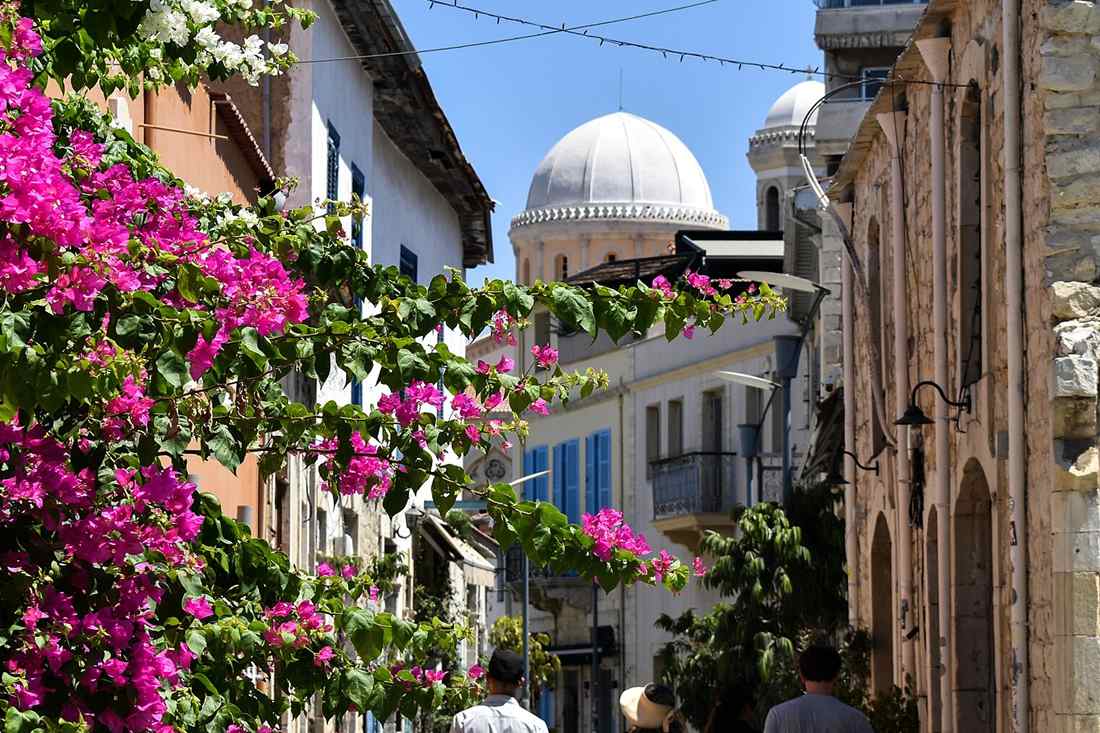 Narrow Limassol old town street with pink bougainvillea, white and stone houses, blue shutters and a white church dome under a blue sky, with visitors walking ahead