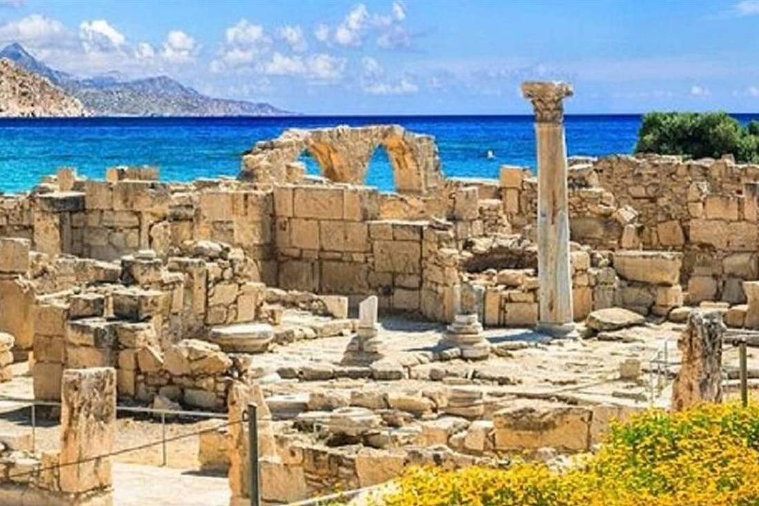 Ruined stone walls, a standing column and arches of an ancient site beside the bright blue sea, with yellow flowers in the foreground