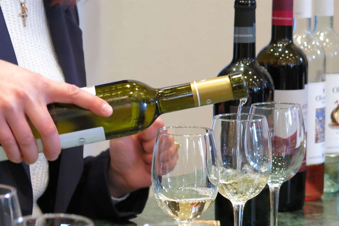 A hand pours white wine from a bottle into a glass at a wine tasting, with bottles of red, rose and white wine standing behind it