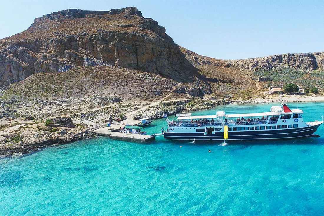 A rocky hill topped with old fortress walls above turquoise water, with a two-deck tour boat moored at a stone jetty at Gramvousa.