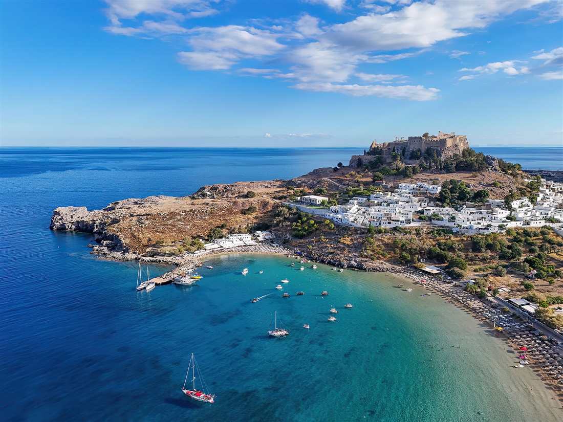 Lindos village and its acropolis on the rock above a curved sandy bay with boats on turquoise water, Rhodes.