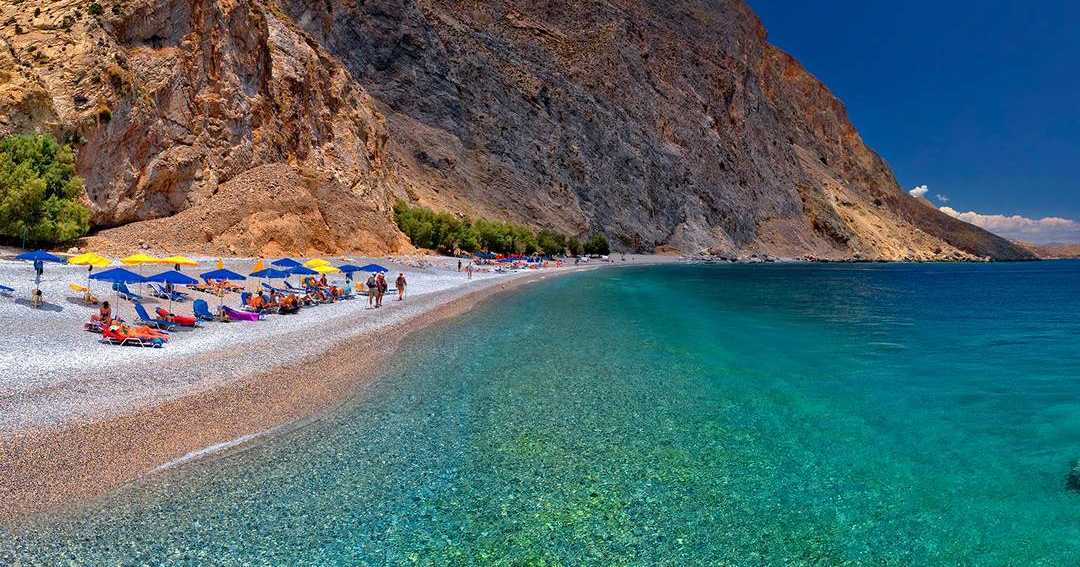 Sweet Water Beach near Loutro, a long pebble shore with umbrellas under steep cliffs and clear turquoise water.