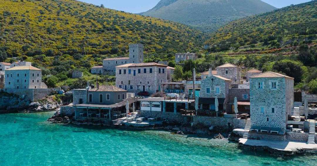 Stone tower houses at the edge of turquoise water in Limeni bay, with green hills and a mountain behind.