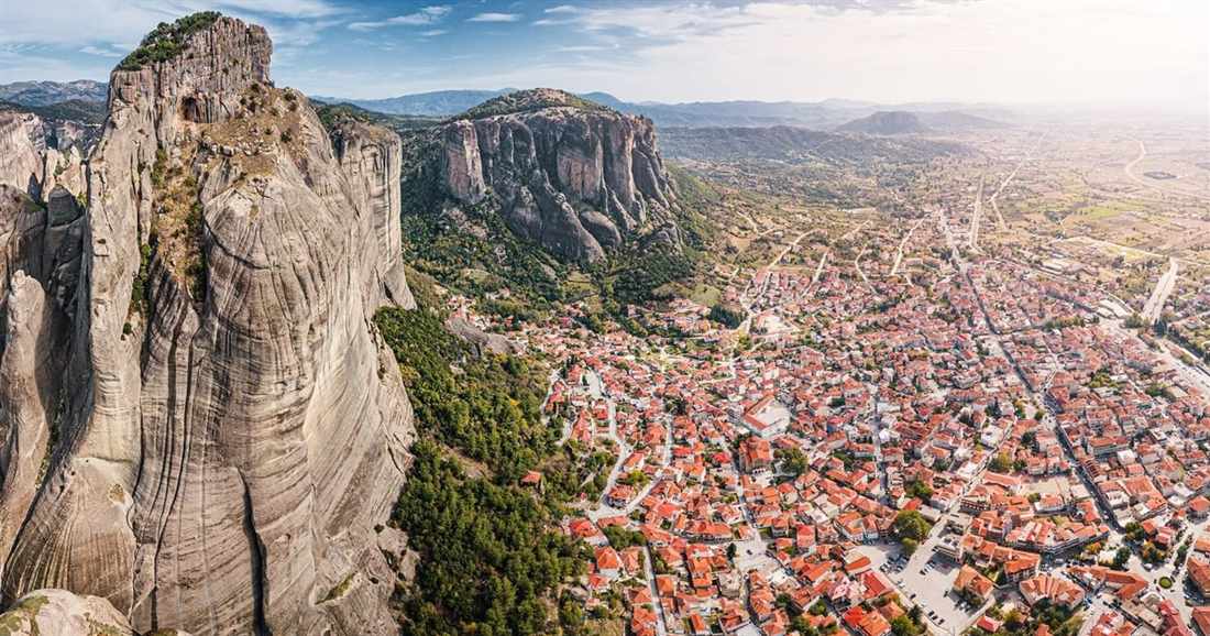 An aerial view of the red roofs of Kalambaka spreading below a tall grey rock pillar at Meteora.