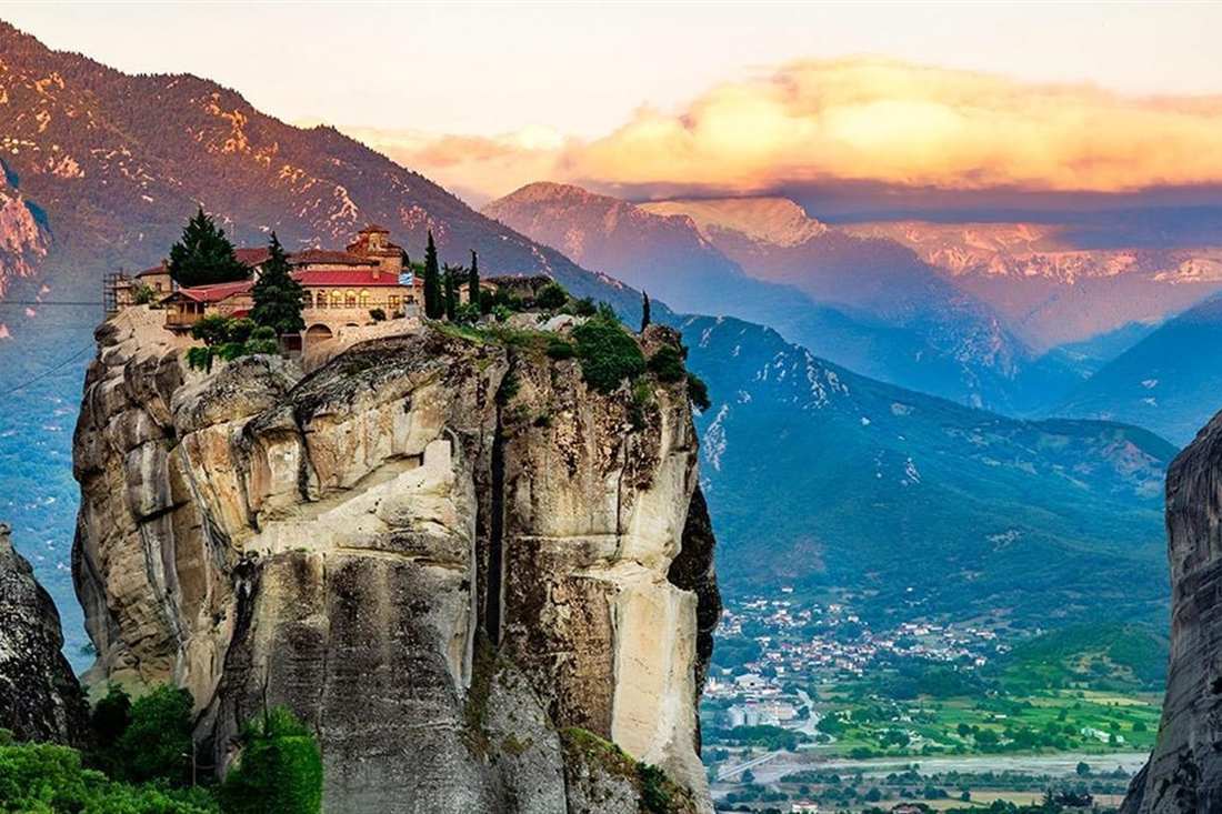 A monastery with red roofs sits on top of a sheer rock pillar at Meteora, with mountains and a pink sunset sky behind.
