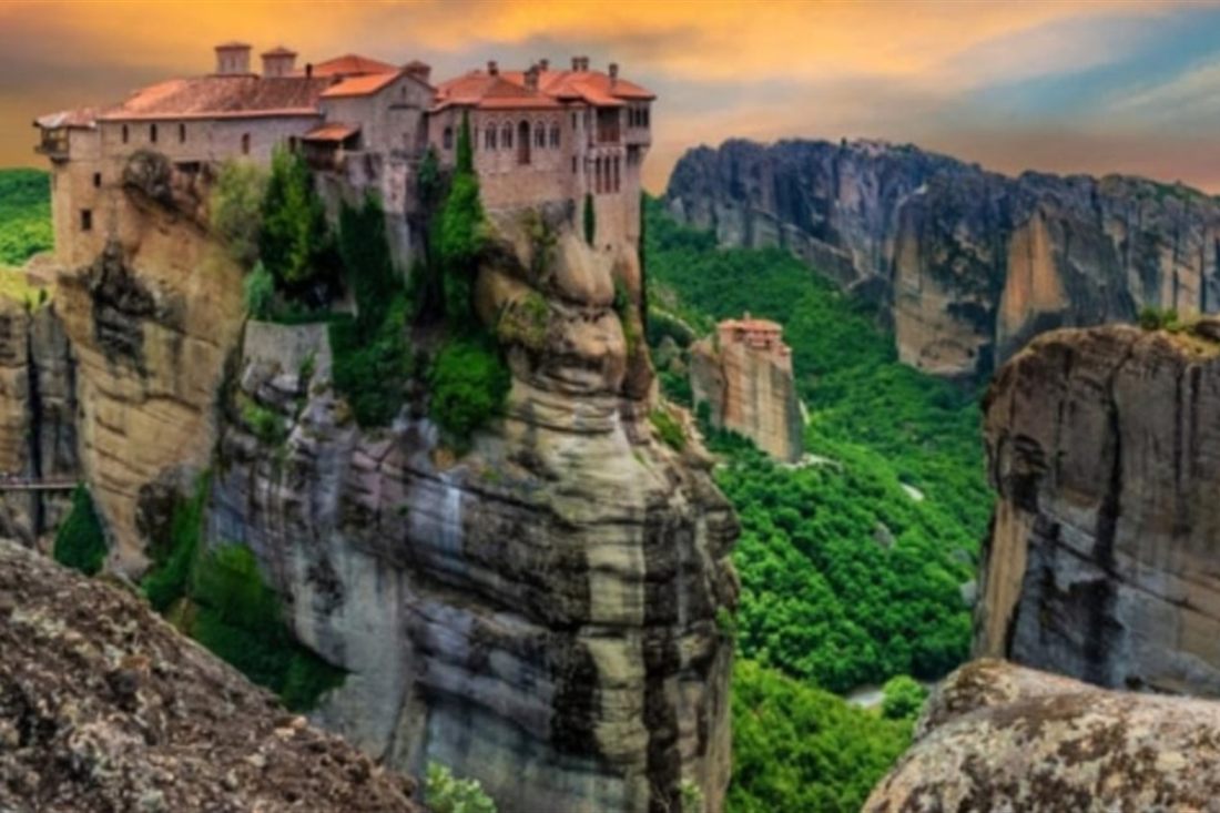 A red-roofed monastery stands on top of a sheer rock pillar at Meteora under a warm evening sky, with green slopes below.