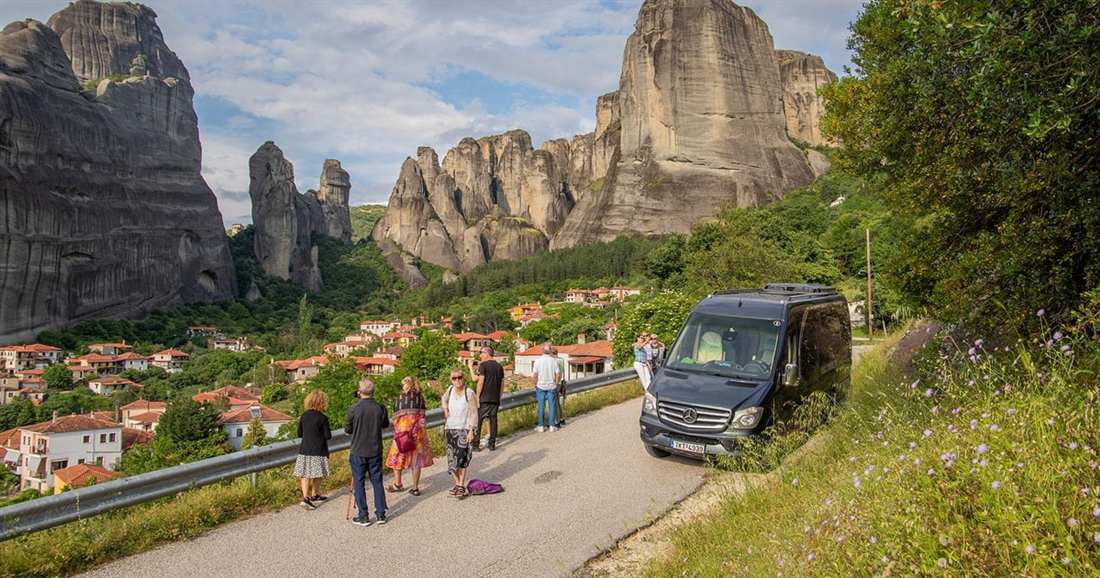 A tour group stands beside a black minibus on a road below the rock pillars of Meteora, with red-roofed houses on the slope.