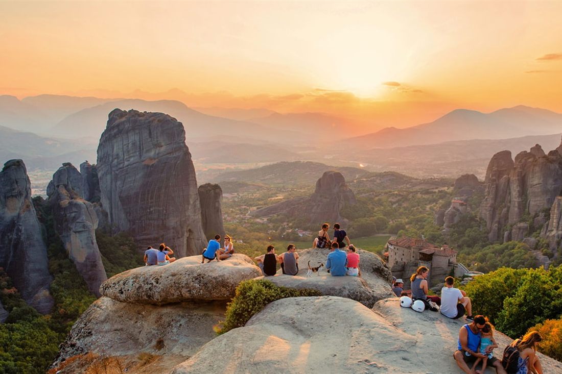 Visitors stand on a rock ledge at sunset above the stone pillars of Meteora, with green valleys and mountains behind.