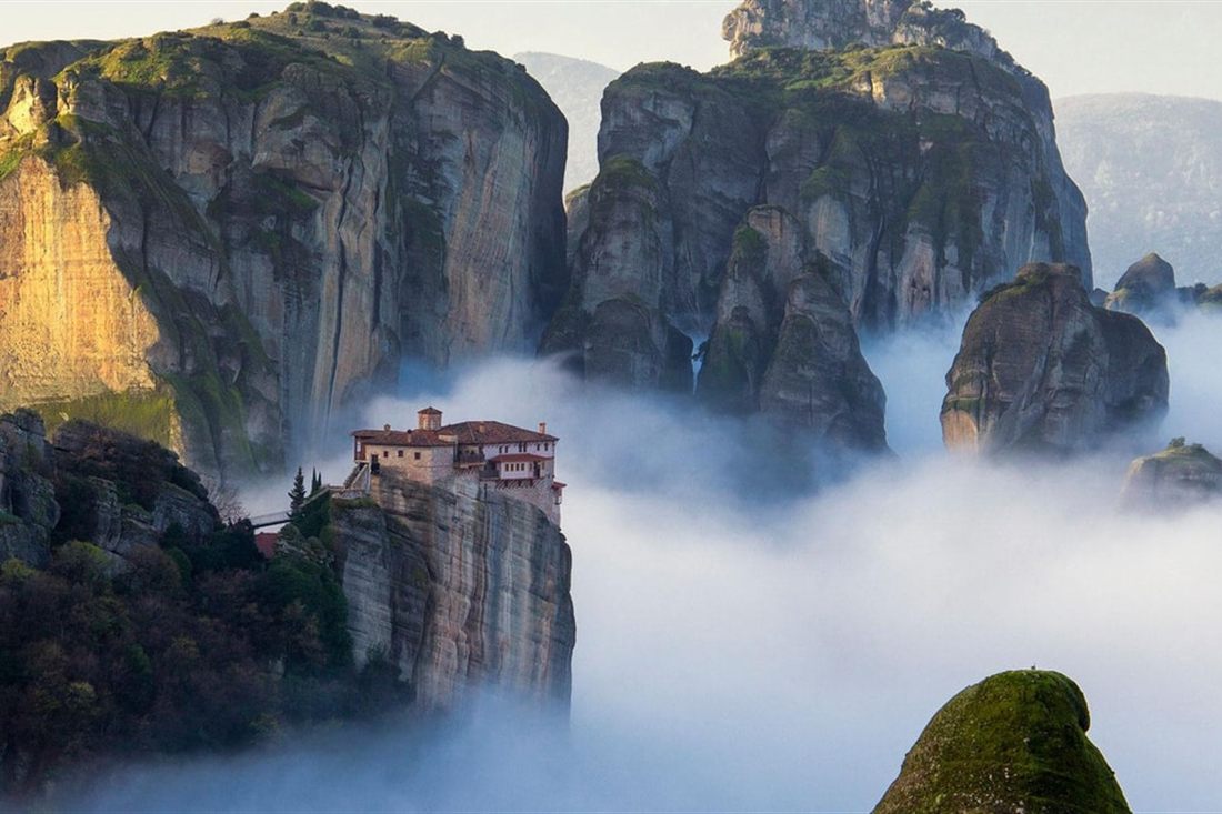 A red-roofed monastery sits on a rock pillar above a sea of white cloud at Meteora, with grey cliffs behind.