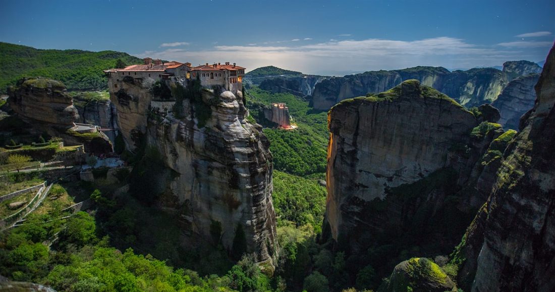 A monastery with red roofs sits on top of a sheer rock pillar at Meteora, above green wooded slopes.