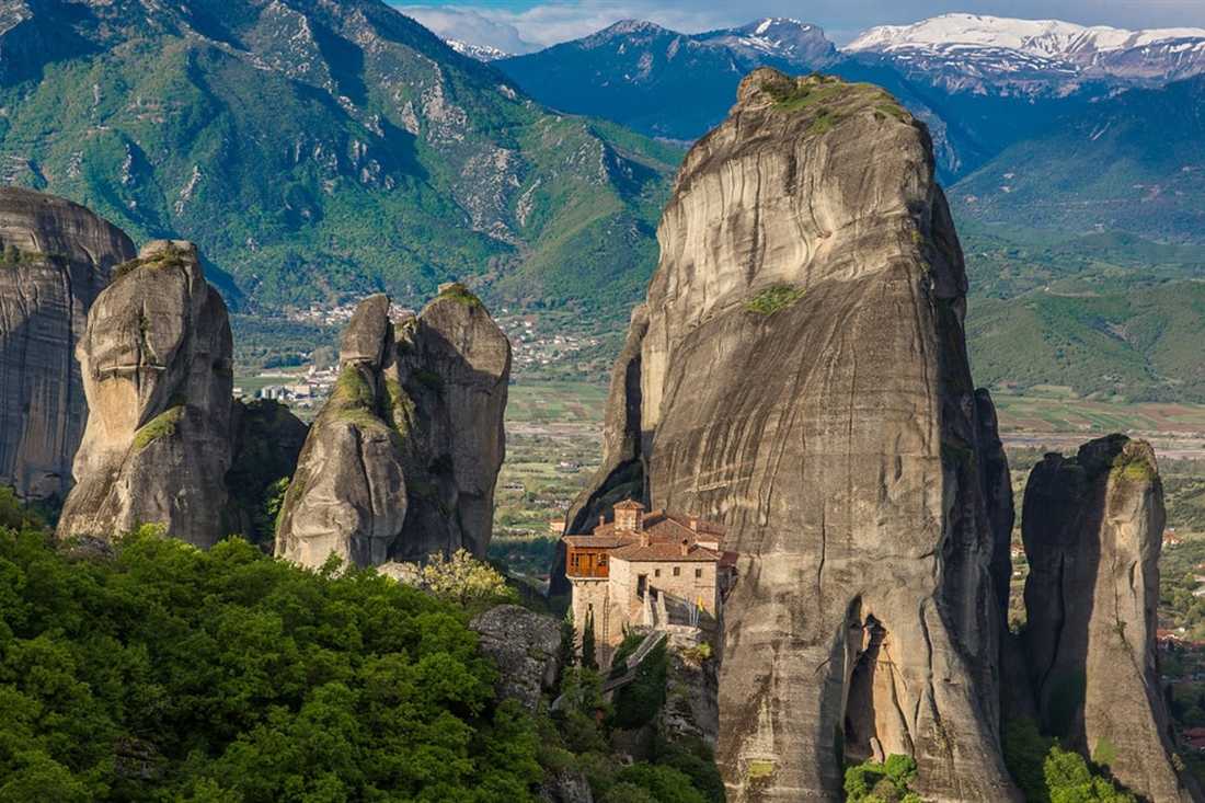 Tall grey rock pillars rise above green forest at Meteora, with a red-roofed monastery at their foot and snowy mountains behind.