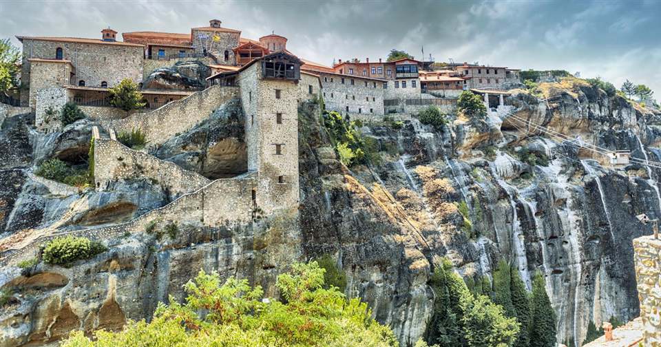 A Meteora monastery with red-tiled roofs on a grey sandstone cliff, with stairs cut into the rock below.