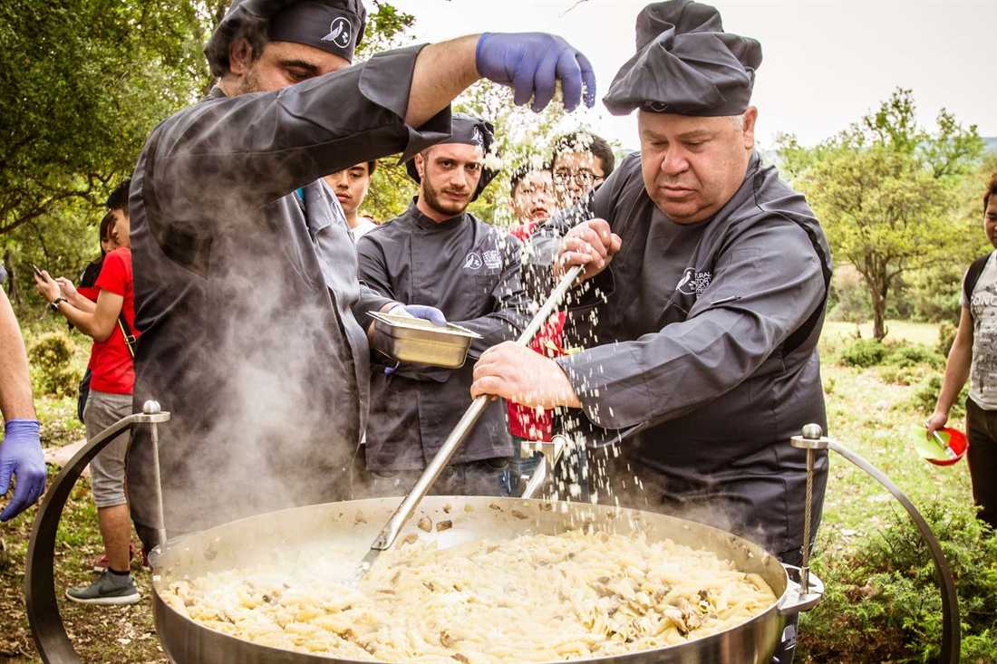 Cooks stirring a large pan of truffle pasta outdoors in the woods below Meteora