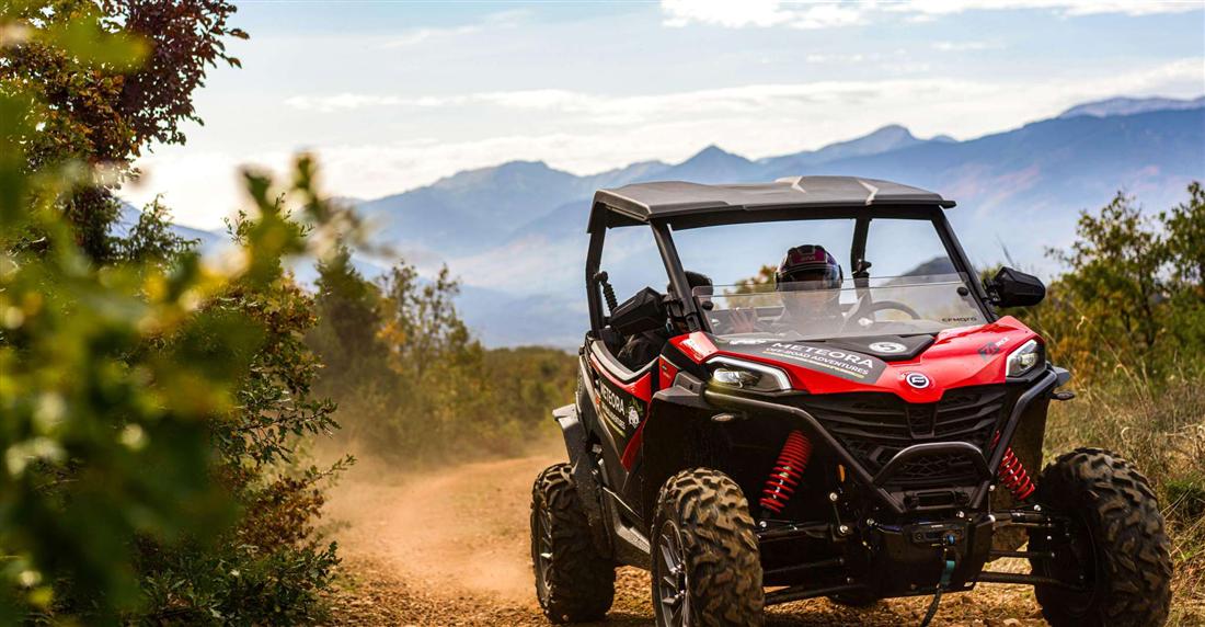 A red off-road buggy with a helmeted driver on a dirt track, with the mountains around Meteora behind