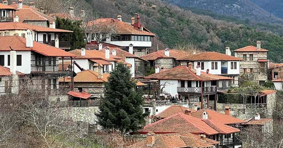 Stone mansions with wooden balconies and red tiled roofs in Metsovo, with a fir tree and mountains behind.