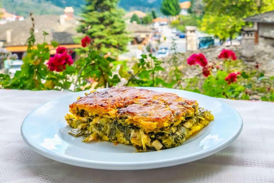 A slice of Greek pie on a white plate with red geraniums and the stone houses of Metsovo behind