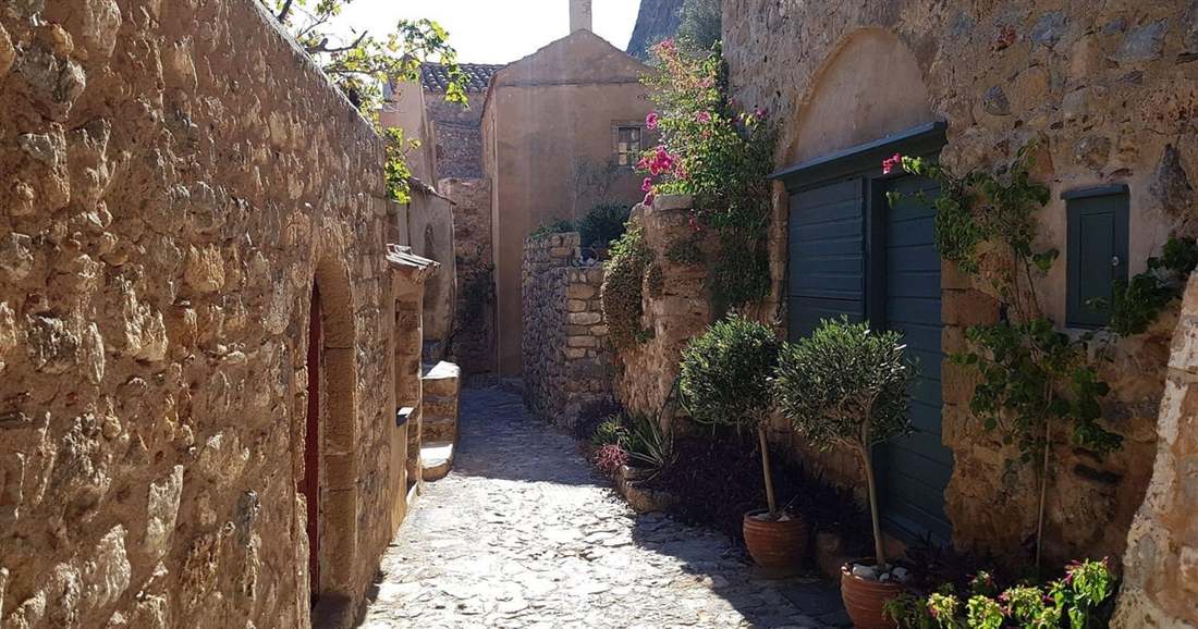 A narrow cobbled lane between old stone houses in the Lower Town of Monemvasia, with potted plants by the doors.