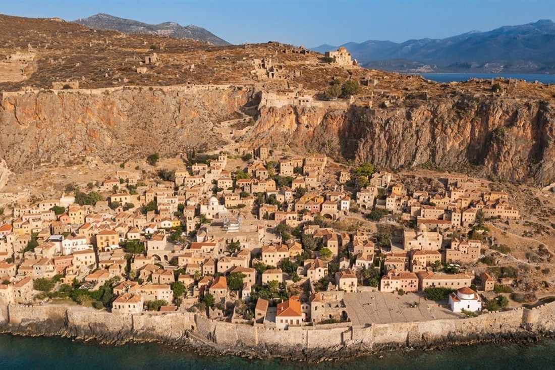 Aerial view of the Lower Town of Monemvasia below the great rock, with stone houses, sea walls and blue water.