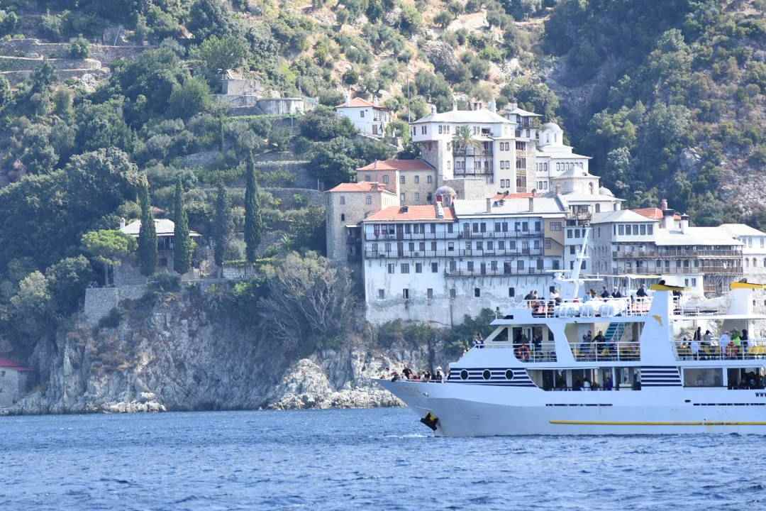 A white cruise boat sailing past a large monastery on the green, rocky west coast of Mount Athos.