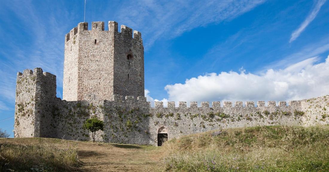 The stone keep and battlemented walls of Platamon castle on a grassy hilltop under a blue sky.