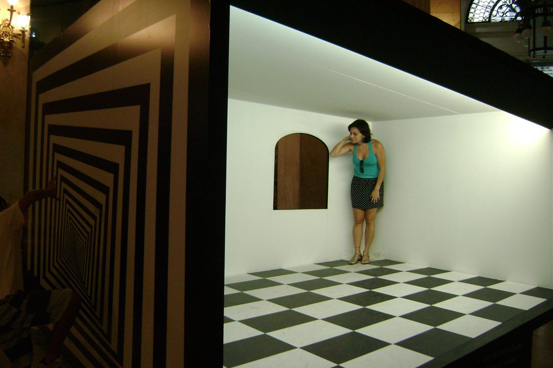 A visitor inside an Ames room, a tilted room that makes people look giant or tiny, the kind of exhibit seen at the Museum of Illusions Thessaloniki.