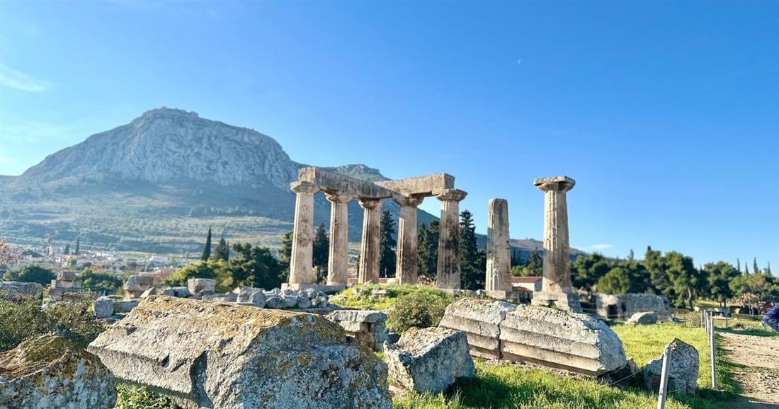 The Doric columns of the Temple of Apollo at Ancient Corinth among ruins, with the rock of Acrocorinth behind under a blue sky.