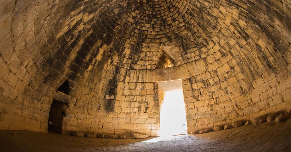 The inside of the Treasury of Atreus at Mycenae, a stone beehive dome with daylight coming through the tall doorway.