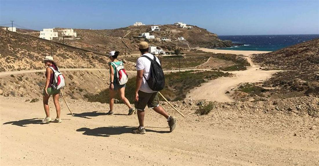 Hikers with poles walking a dirt track above a bay on the Mykonos coast.