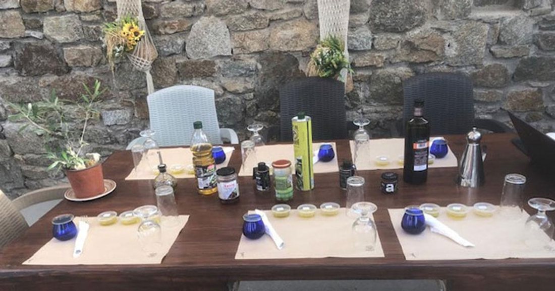 A long table set with blue tasting glasses, bottles of olive oil and paper mats in a stone farmyard, ready for a Mykonos olive oil tasting.